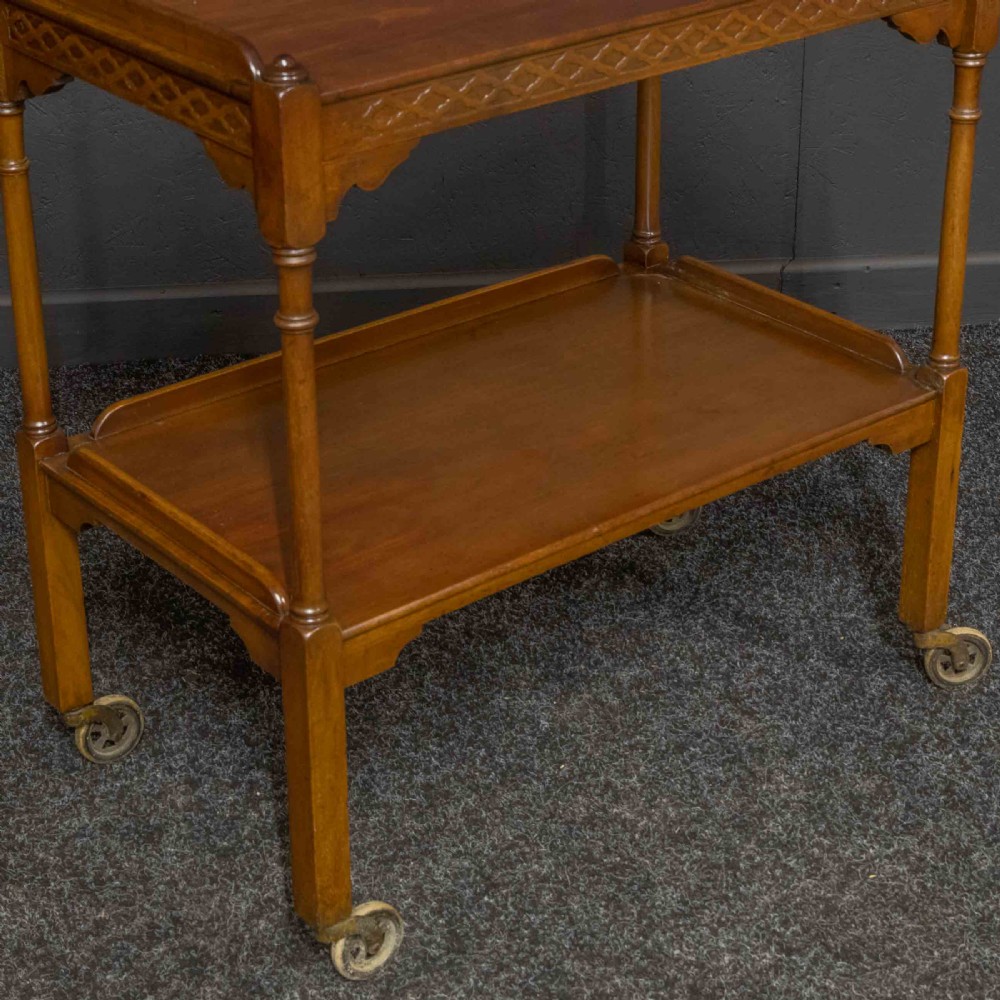 A two-tiered warm brown polished wooden tea trolley featuring a carved geometric frieze, turned column supports, gallery edges, and four small metal castors. View III