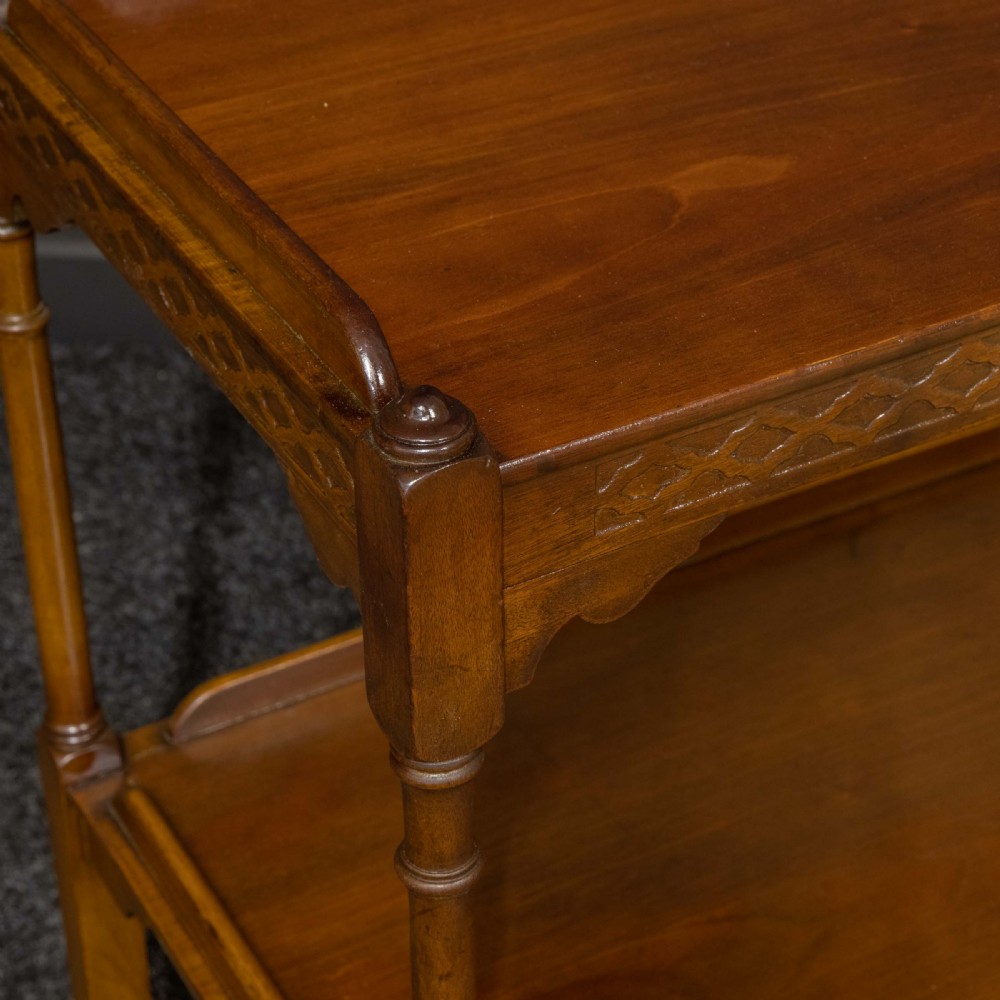 A two-tiered warm brown polished wooden tea trolley featuring a carved geometric frieze, turned column supports, gallery edges, and four small metal castors. View IV