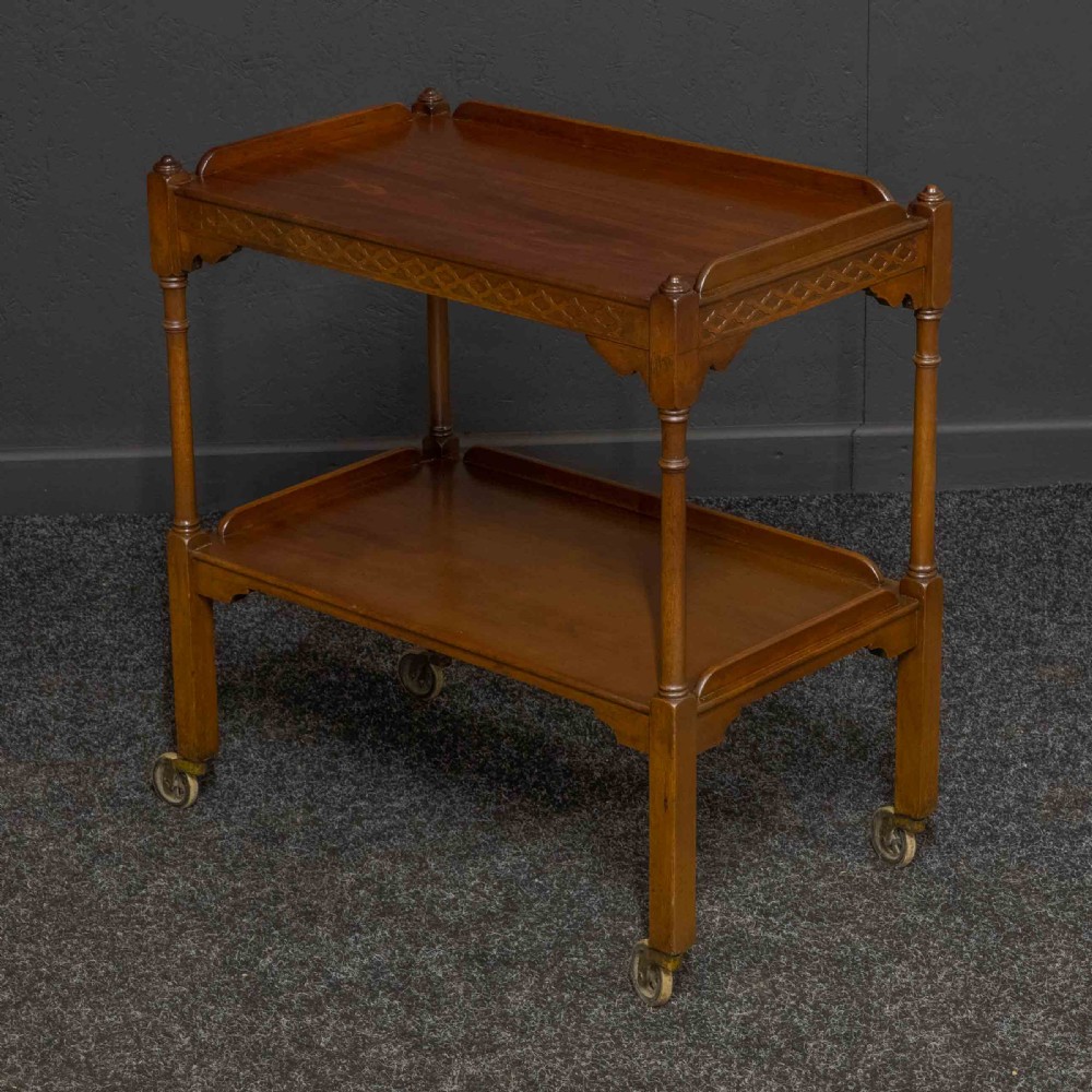 A two-tiered warm brown polished wooden tea trolley featuring a carved geometric frieze, turned column supports, gallery edges, and four small metal castors. View V