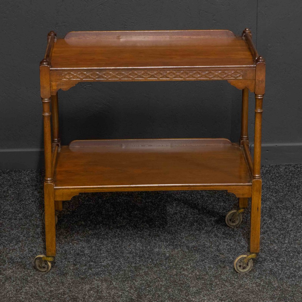 A two-tiered warm brown polished wooden tea trolley featuring a carved geometric frieze, turned column supports, gallery edges, and four small metal castors. View VI