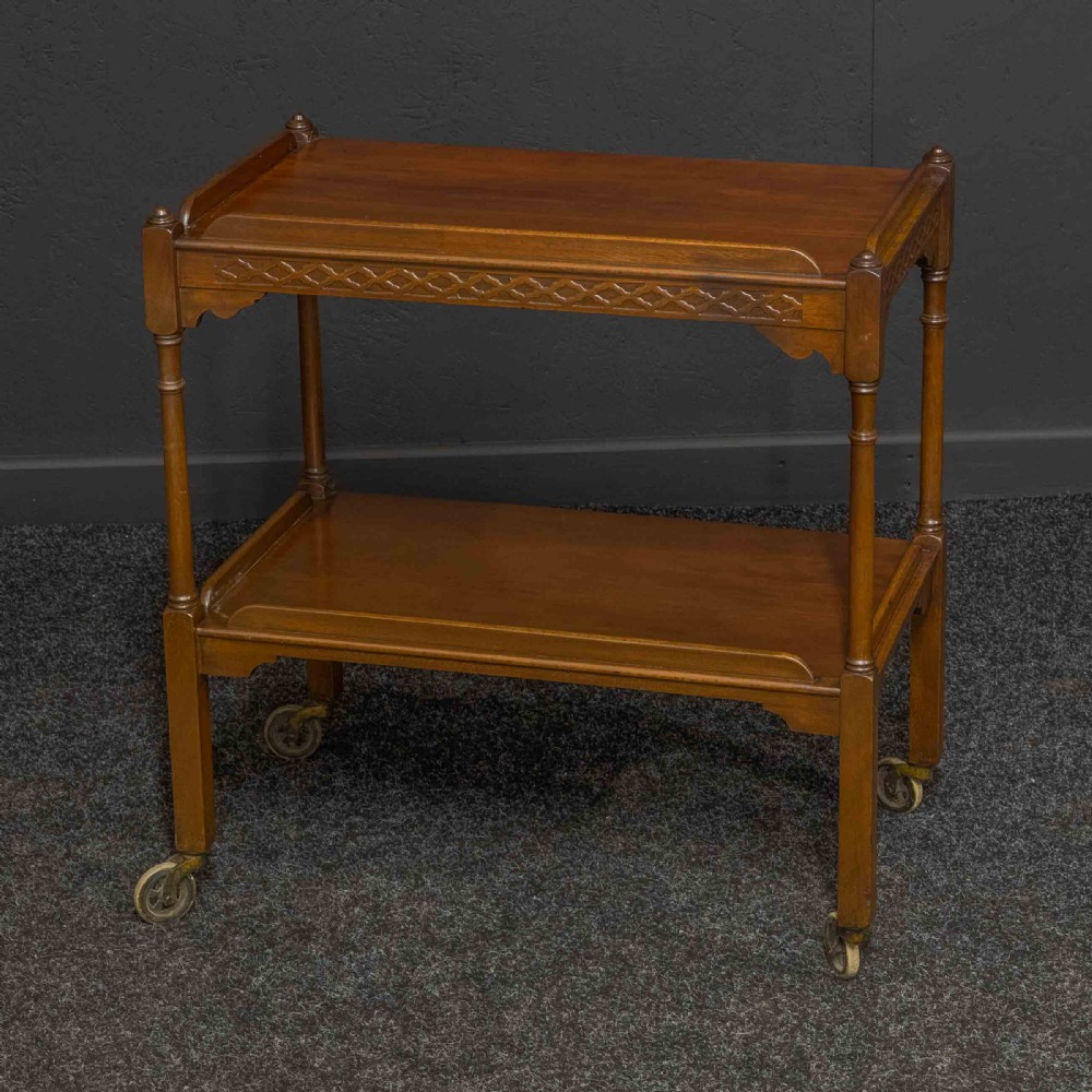 A two-tiered warm brown polished wooden tea trolley featuring a carved geometric frieze, turned column supports, gallery edges, and four small metal castors. View VII
