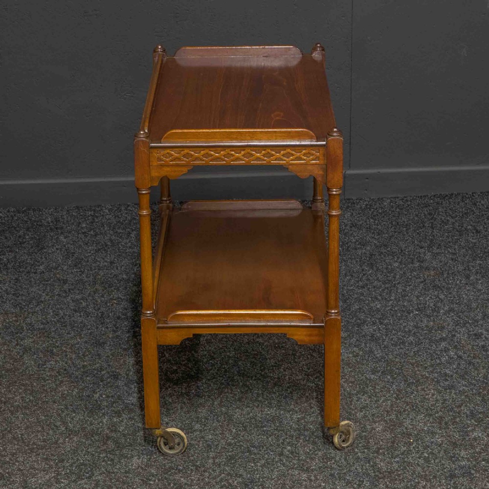 A two-tiered warm brown polished wooden tea trolley featuring a carved geometric frieze, turned column supports, gallery edges, and four small metal castors. View VIII