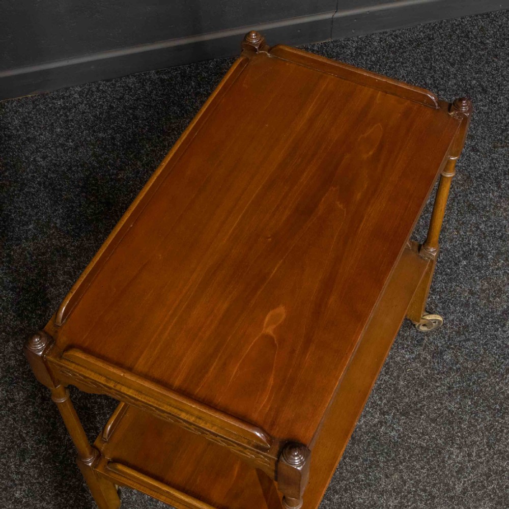 A two-tiered warm brown polished wooden tea trolley featuring a carved geometric frieze, turned column supports, gallery edges, and four small metal castors. View IX