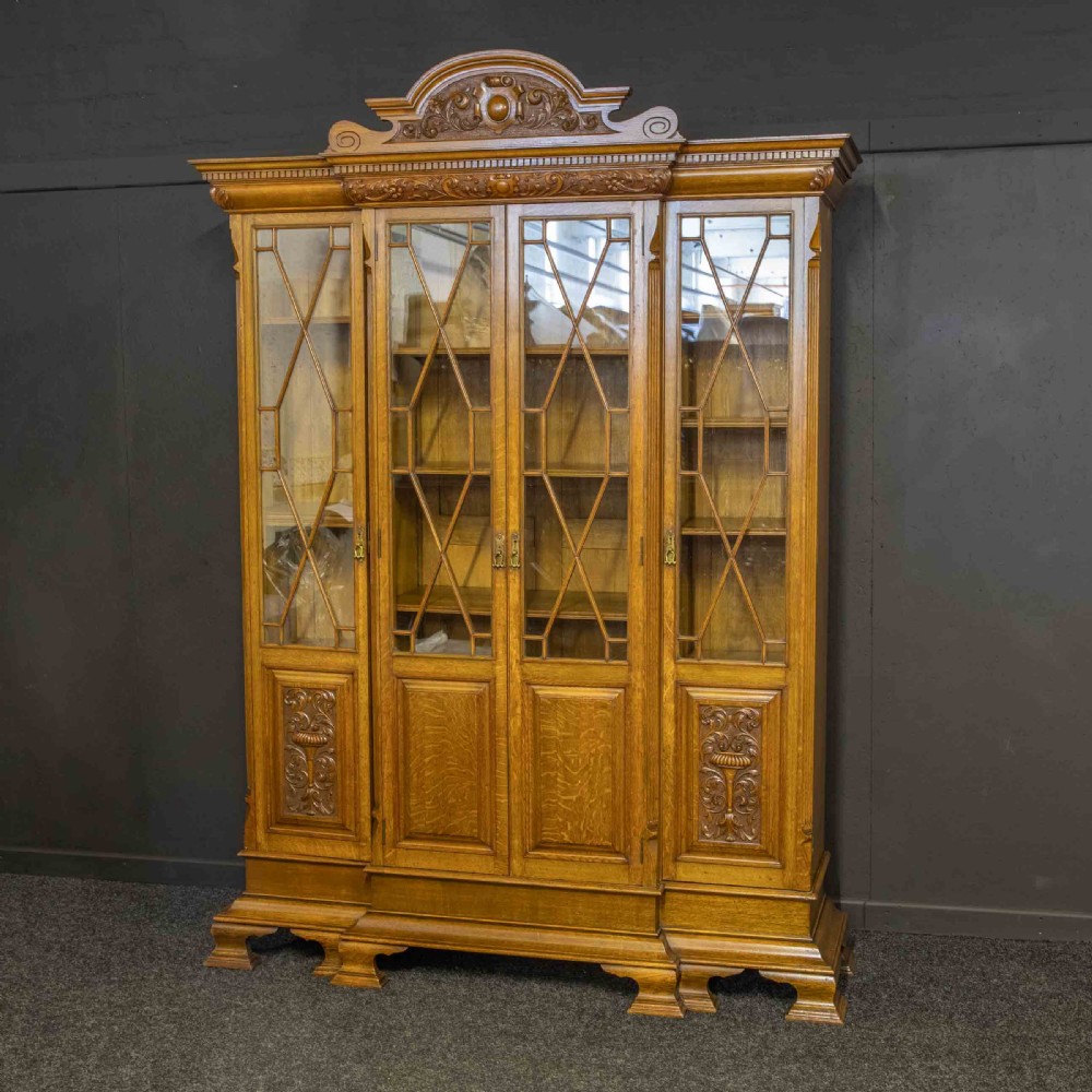 An Edwardian oak breakfront bookcase featuring four glazed upper doors with geometric astragal bars, carved lower panel doors, and a decorative carved top pediment.