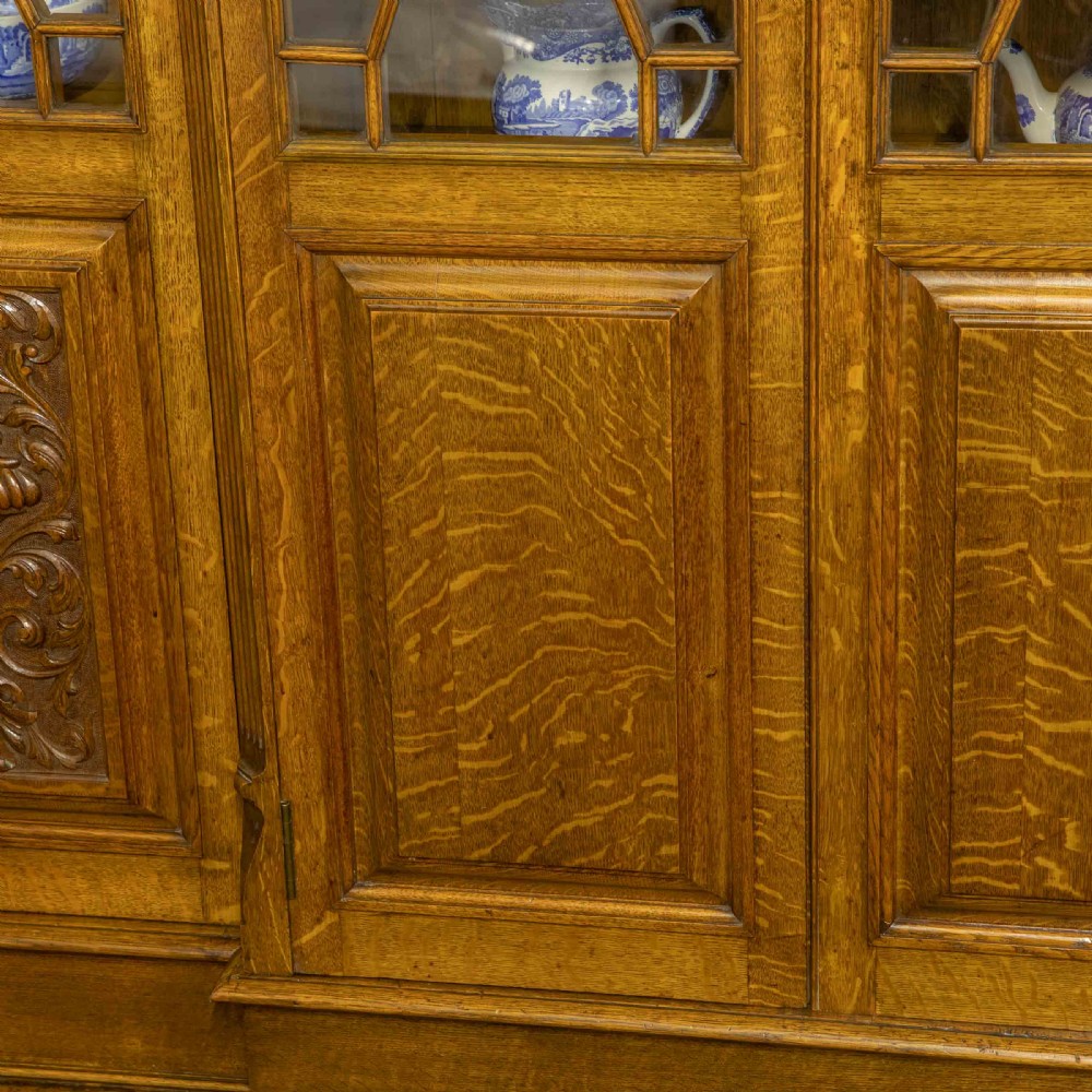 An Edwardian oak breakfront bookcase featuring four glazed upper doors with geometric astragal bars, carved lower panel doors, and a decorative carved top pediment. View II