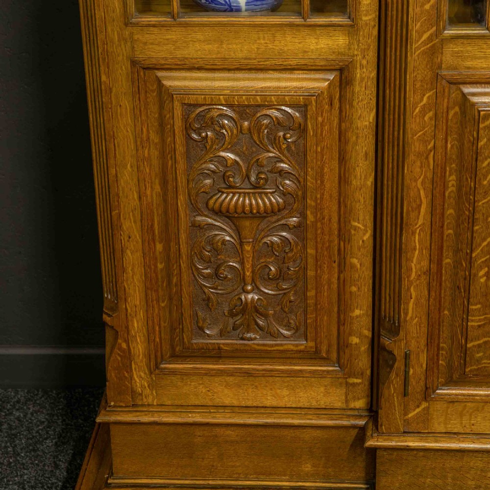An Edwardian oak breakfront bookcase featuring four glazed upper doors with geometric astragal bars, carved lower panel doors, and a decorative carved top pediment. View III