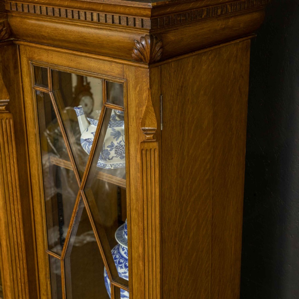 An Edwardian oak breakfront bookcase featuring four glazed upper doors with geometric astragal bars, carved lower panel doors, and a decorative carved top pediment. View VI