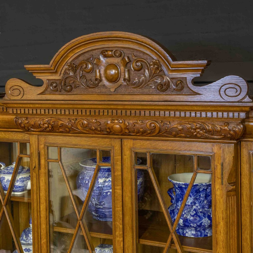 An Edwardian oak breakfront bookcase featuring four glazed upper doors with geometric astragal bars, carved lower panel doors, and a decorative carved top pediment. View VII