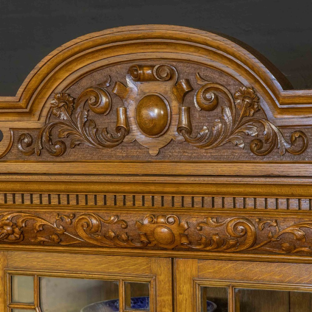 An Edwardian oak breakfront bookcase featuring four glazed upper doors with geometric astragal bars, carved lower panel doors, and a decorative carved top pediment. View VIII