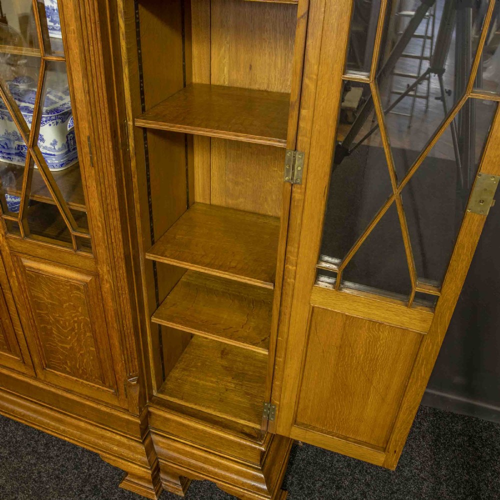 An Edwardian oak breakfront bookcase featuring four glazed upper doors with geometric astragal bars, carved lower panel doors, and a decorative carved top pediment. View IX