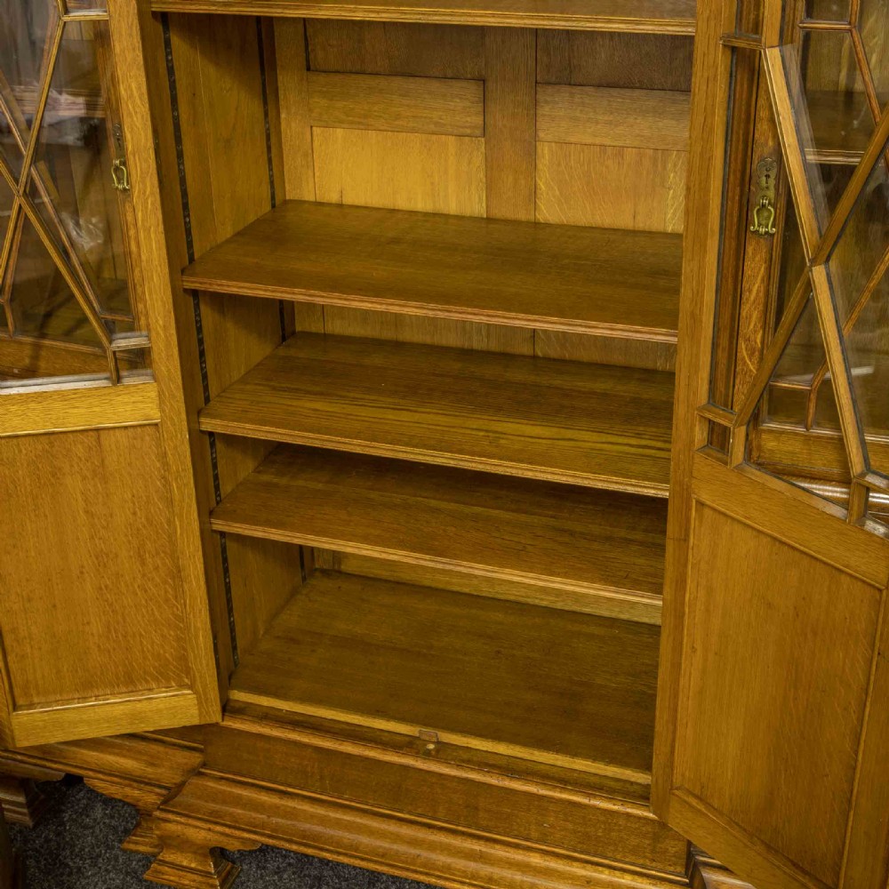 An Edwardian oak breakfront bookcase featuring four glazed upper doors with geometric astragal bars, carved lower panel doors, and a decorative carved top pediment. View X