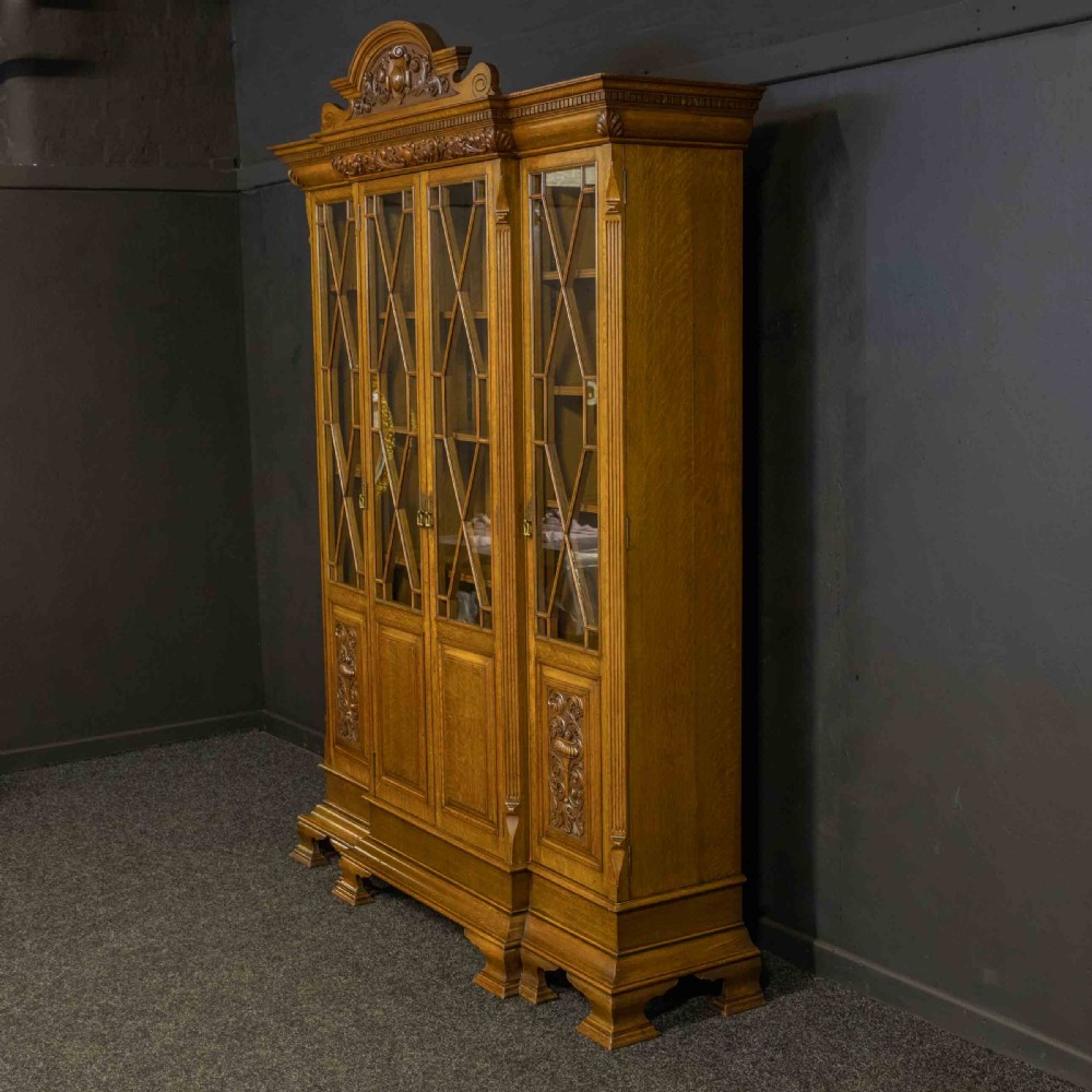 An Edwardian oak breakfront bookcase featuring four glazed upper doors with geometric astragal bars, carved lower panel doors, and a decorative carved top pediment. View XI