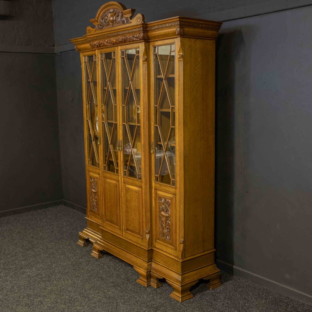 An Edwardian oak breakfront bookcase featuring four glazed upper doors with geometric astragal bars, carved lower panel doors, and a decorative carved top pediment. View XII