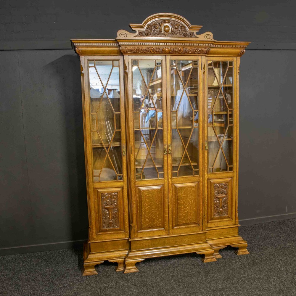 An Edwardian oak breakfront bookcase featuring four glazed upper doors with geometric astragal bars, carved lower panel doors, and a decorative carved top pediment. View XIII