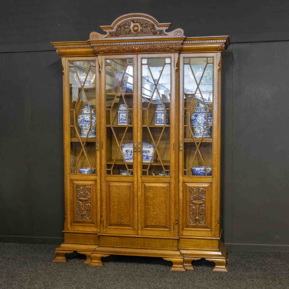 An Edwardian oak breakfront bookcase featuring four glazed upper doors with geometric astragal bars, carved lower panel doors, and a decorative carved top pediment. View XV