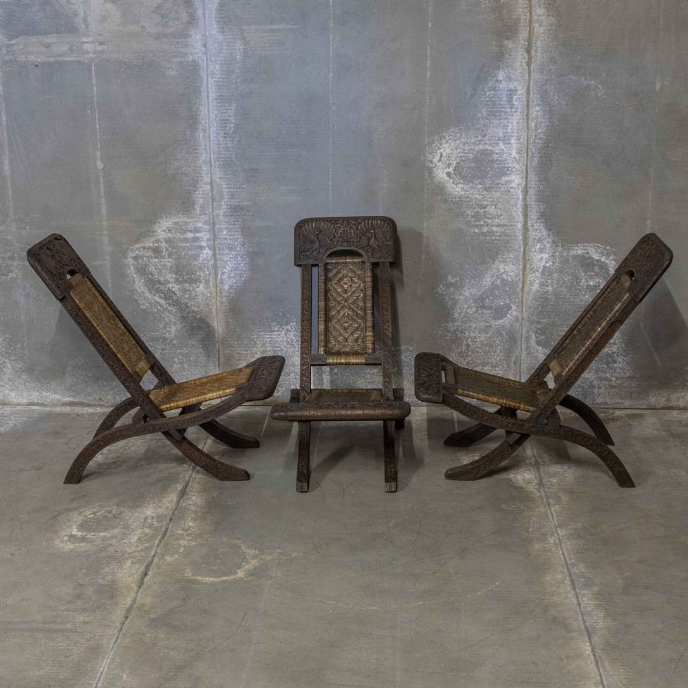 A set of three dark hand-carved wooden folding chairs featuring ornate foliate reliefs and woven rattan seat panels.