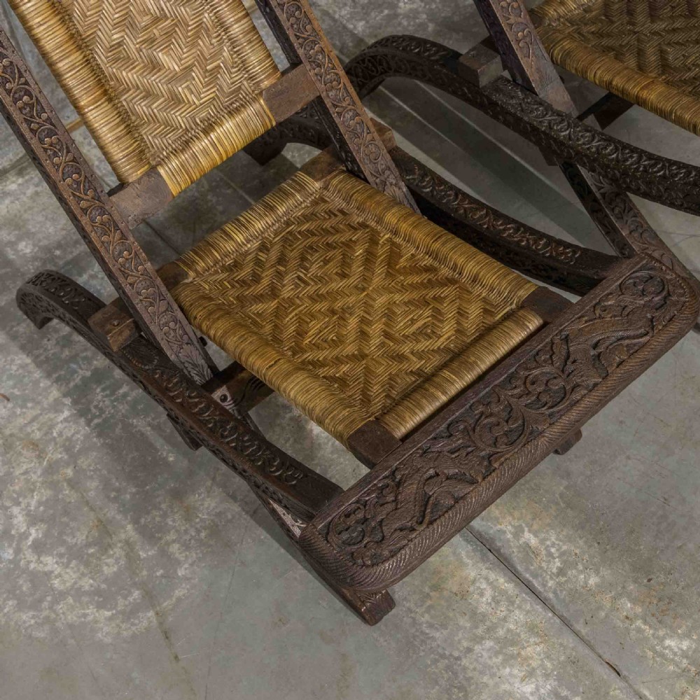 A set of three dark hand-carved wooden folding chairs featuring ornate foliate reliefs and woven rattan seat panels. View VI