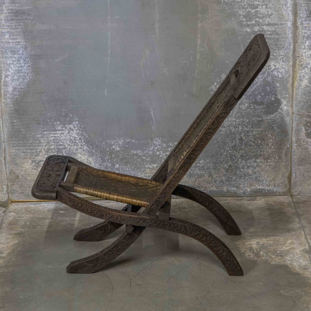 A set of three dark hand-carved wooden folding chairs featuring ornate foliate reliefs and woven rattan seat panels. View X
