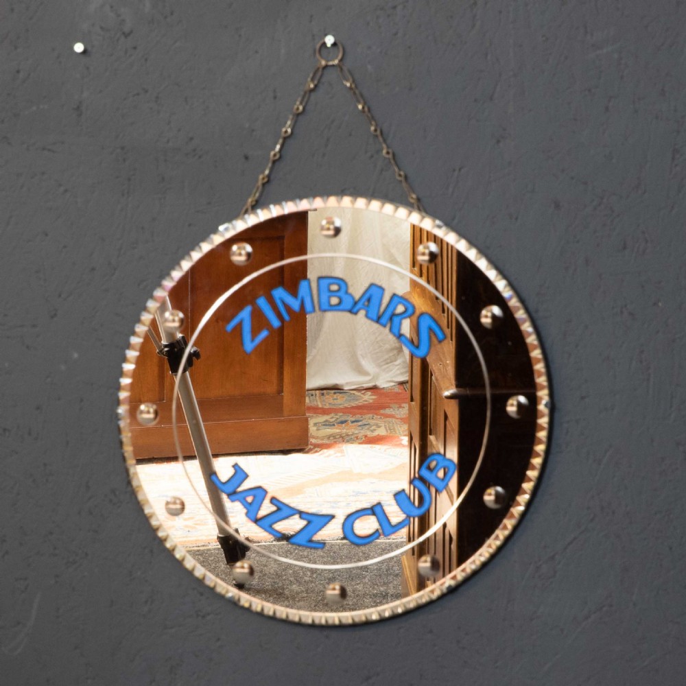 A circular frameless wall mirror featuring blue typography that reads Zimbars Jazz Club, suspended by a top chain. View II