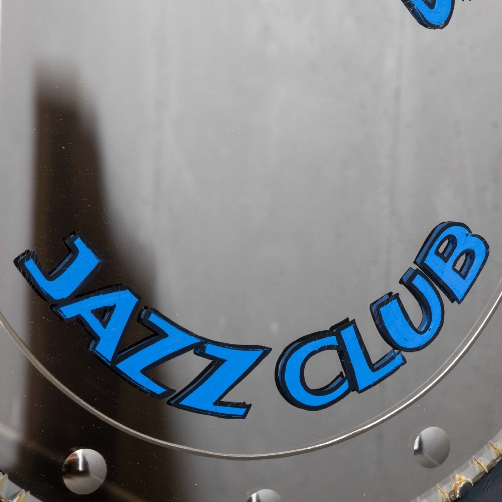 A circular frameless wall mirror featuring blue typography that reads Zimbars Jazz Club, suspended by a top chain. View V
