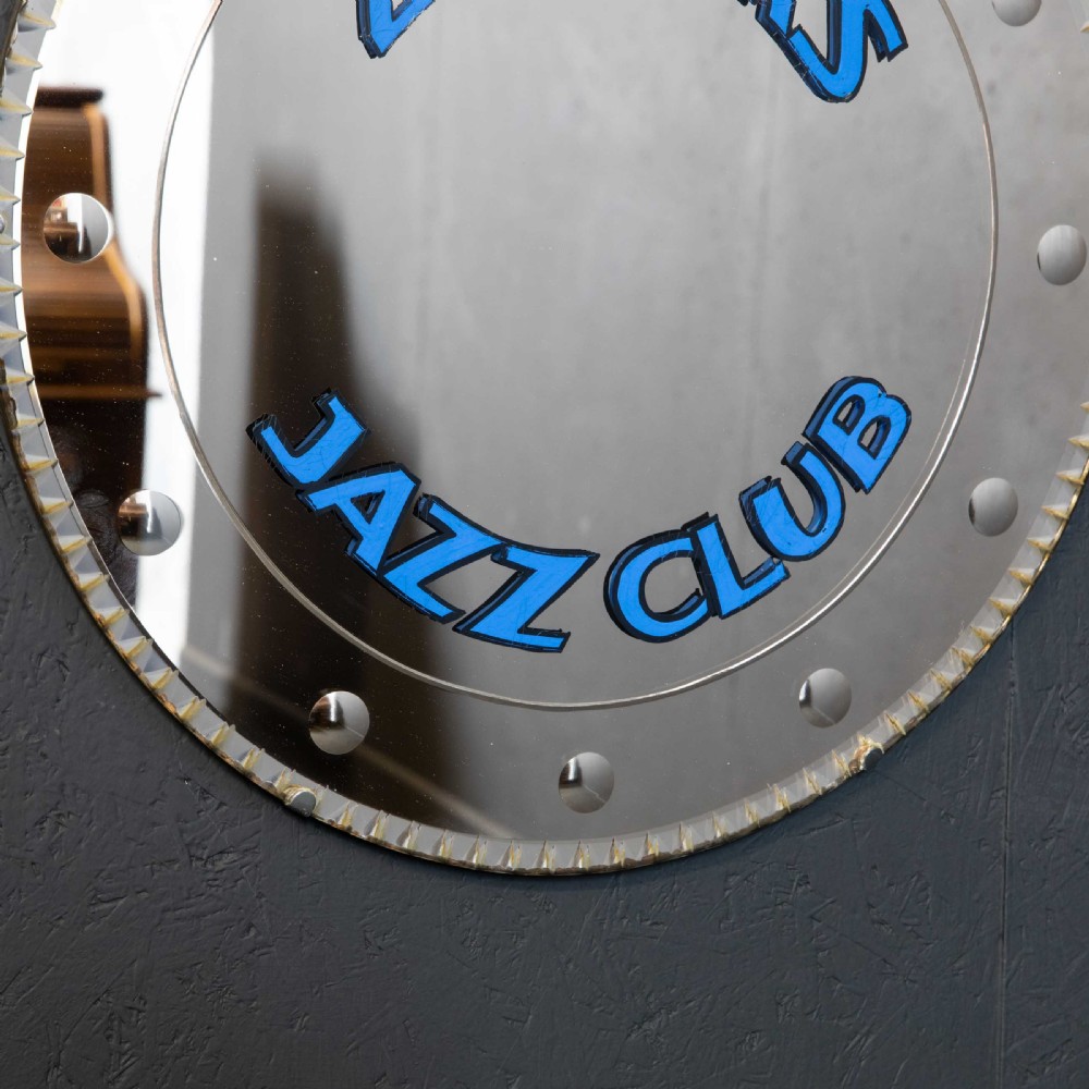 A circular frameless wall mirror featuring blue typography that reads Zimbars Jazz Club, suspended by a top chain. View VII