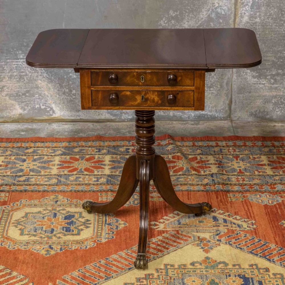 A Regency mahogany drop-leaf worktable featuring two drawers with wooden knobs, a turned pedestal column, and tripod legs with brass paw feet.