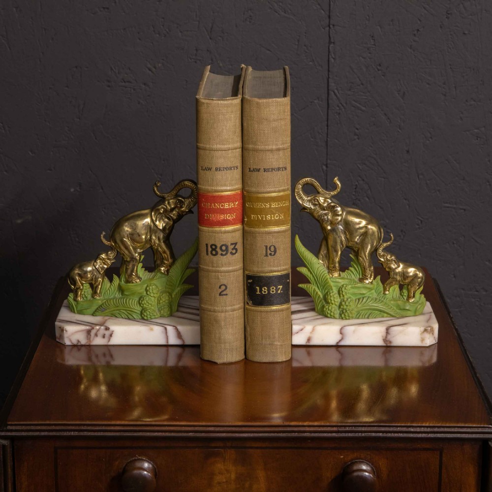 A pair of Art Deco brass elephant bookends with green painted foliage bases mounted on white veined marble plinths holding two books.