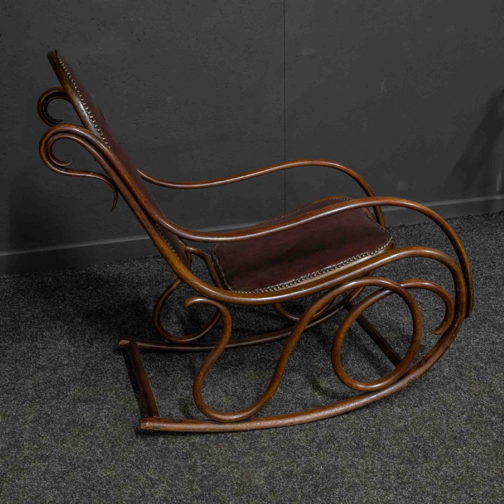 An antique Thonet steam-bent wood rocking chair featuring dark brown leather upholstered seating and brass studding along the frame. View III