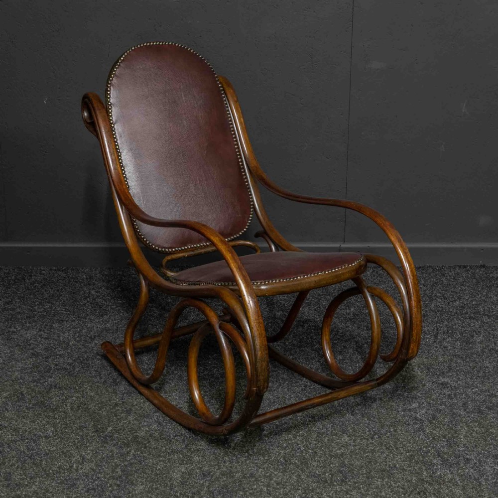 An antique Thonet steam-bent wood rocking chair featuring dark brown leather upholstered seating and brass studding along the frame. View V