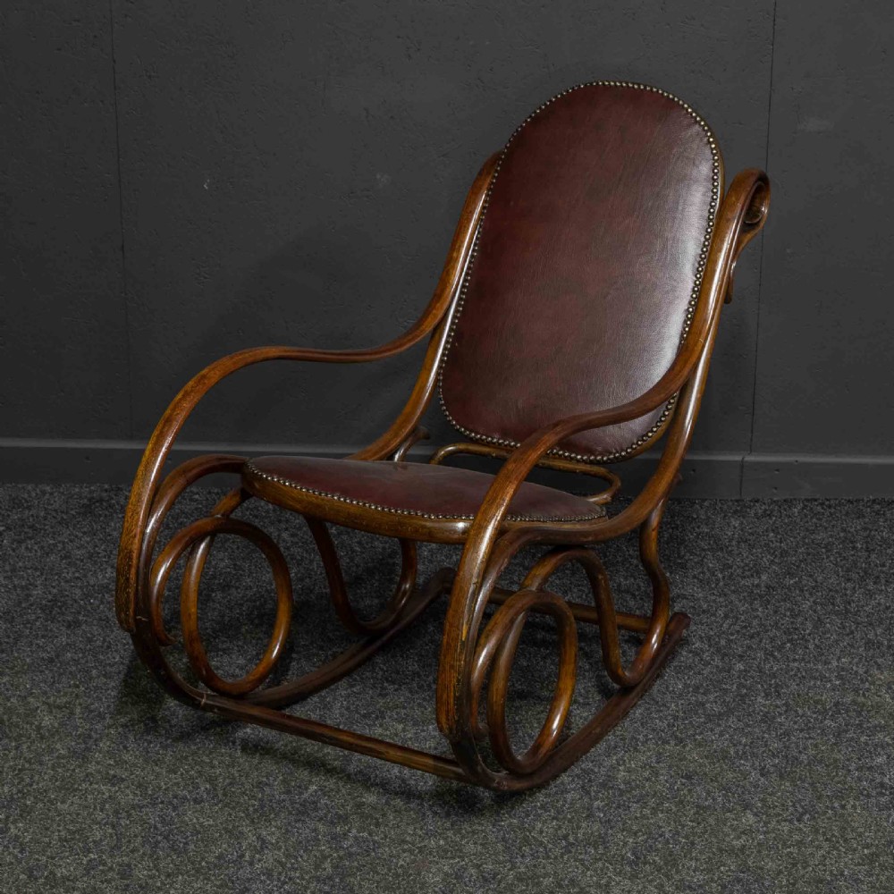 An antique Thonet steam-bent wood rocking chair featuring dark brown leather upholstered seating and brass studding along the frame. View VI