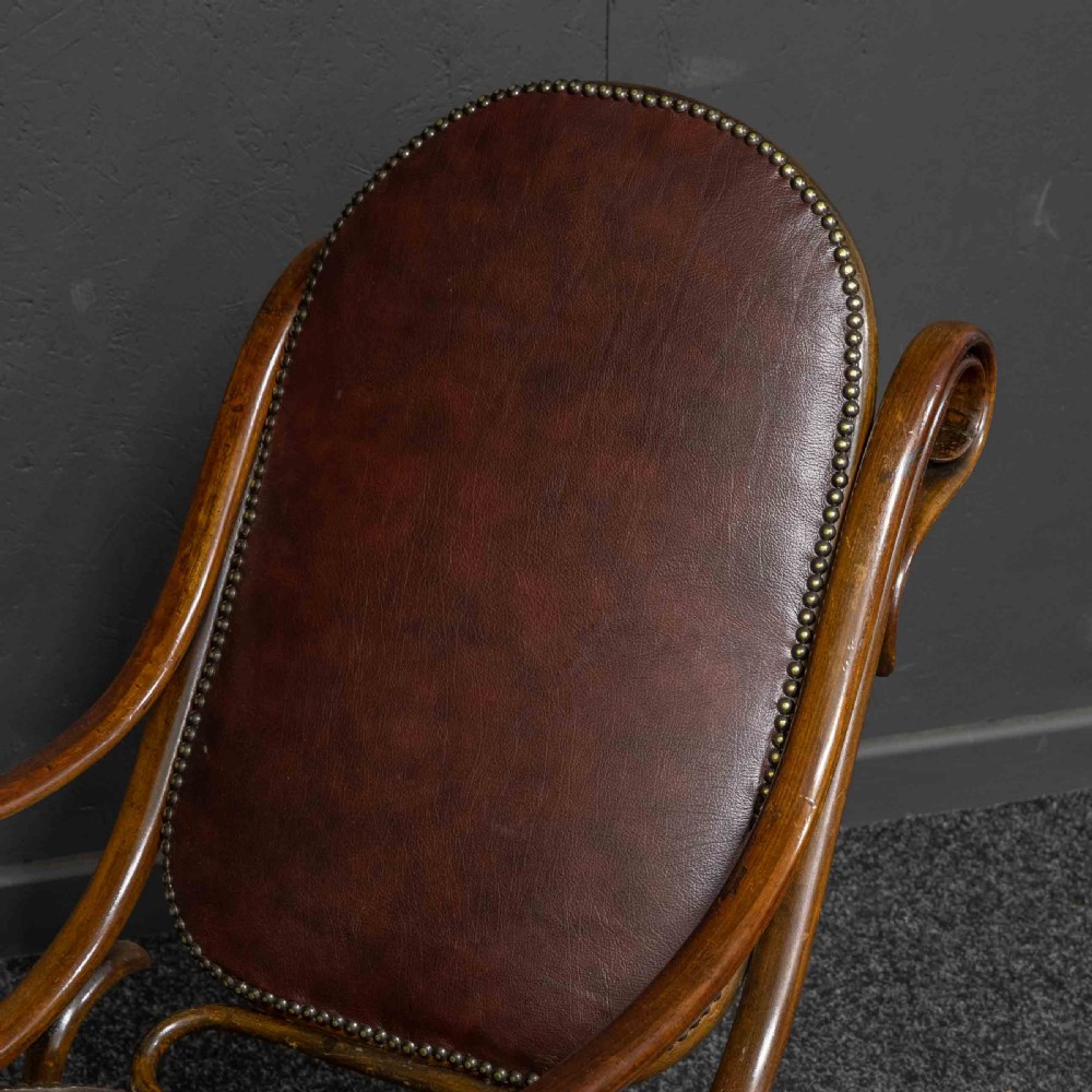 An antique Thonet steam-bent wood rocking chair featuring dark brown leather upholstered seating and brass studding along the frame. View X