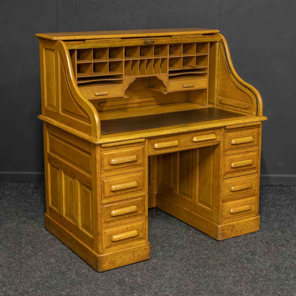 An early 20th-century golden oak roll top desk featuring open fitted pigeonhole compartments, a dark leather writing inset, and twin drawer pedestals.