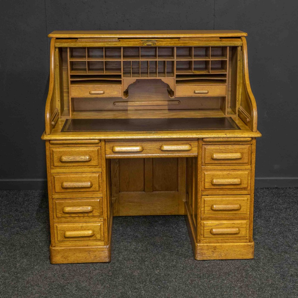 An early 20th-century golden oak roll top desk featuring open fitted pigeonhole compartments, a dark leather writing inset, and twin drawer pedestals. View II