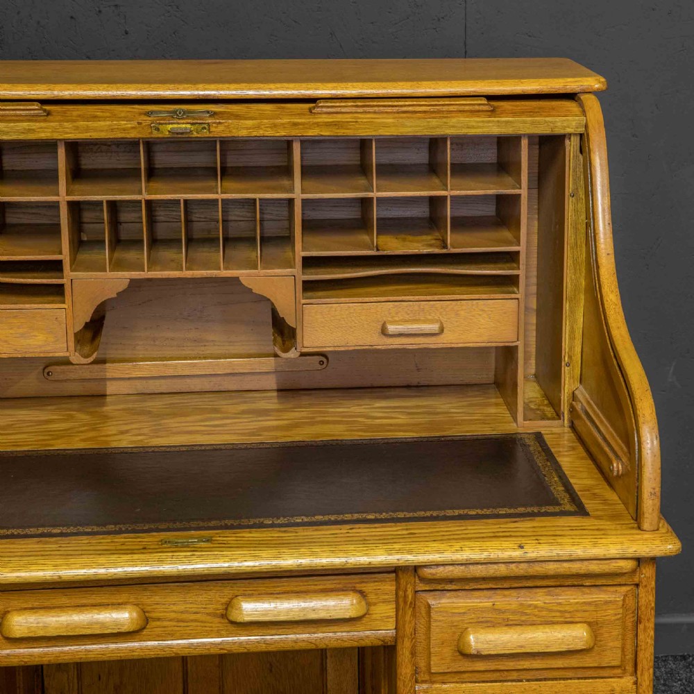 An early 20th-century golden oak roll top desk featuring open fitted pigeonhole compartments, a dark leather writing inset, and twin drawer pedestals. View III