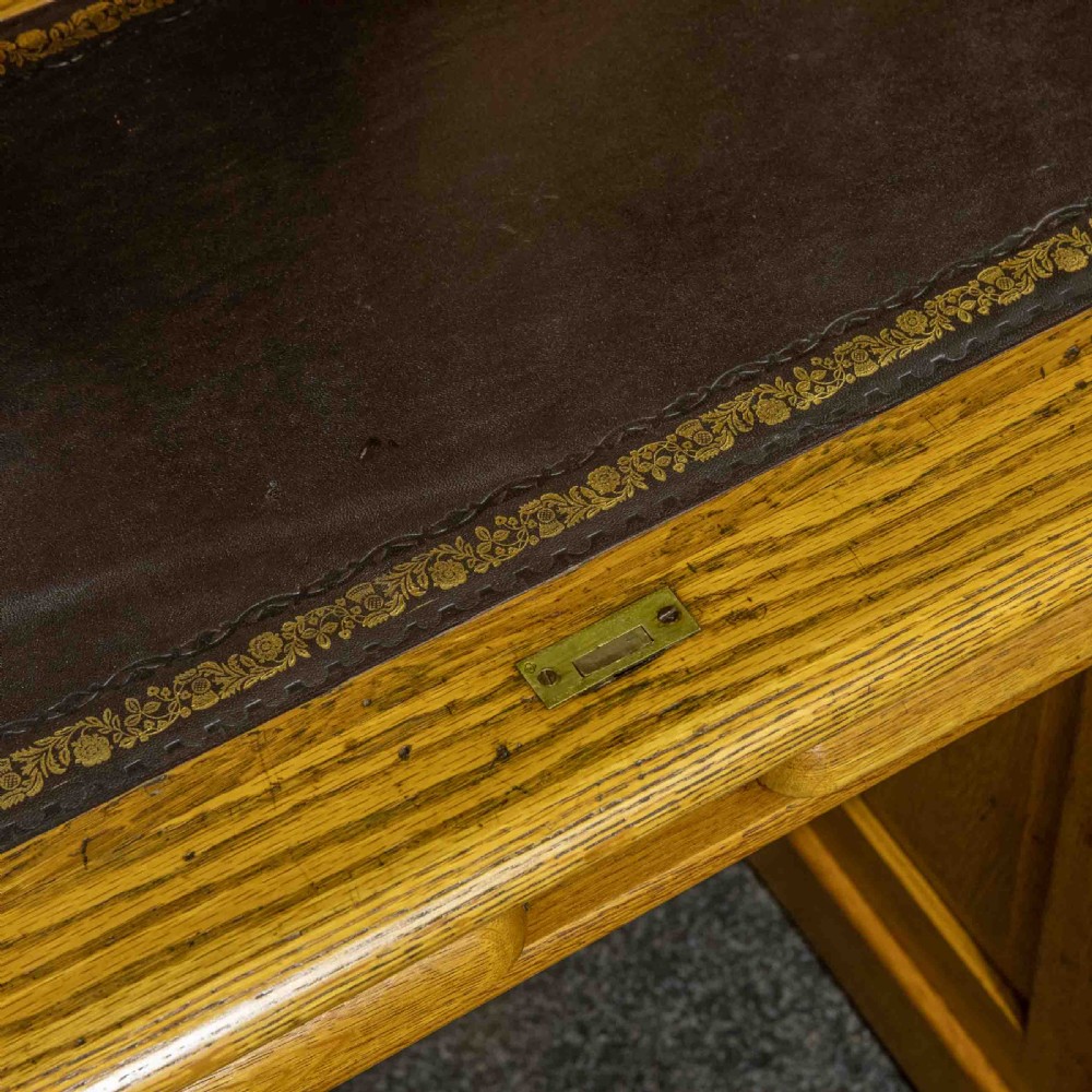 An early 20th-century golden oak roll top desk featuring open fitted pigeonhole compartments, a dark leather writing inset, and twin drawer pedestals. View V