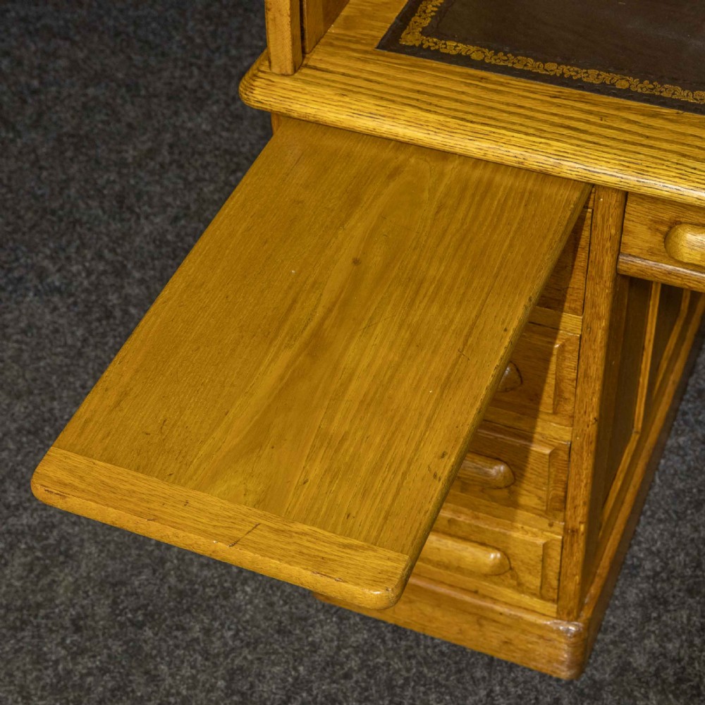 An early 20th-century golden oak roll top desk featuring open fitted pigeonhole compartments, a dark leather writing inset, and twin drawer pedestals. View VI