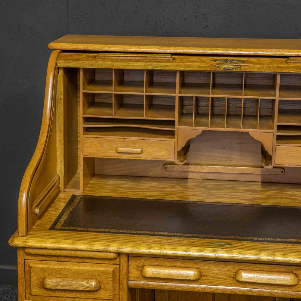 An early 20th-century golden oak roll top desk featuring open fitted pigeonhole compartments, a dark leather writing inset, and twin drawer pedestals. View VII