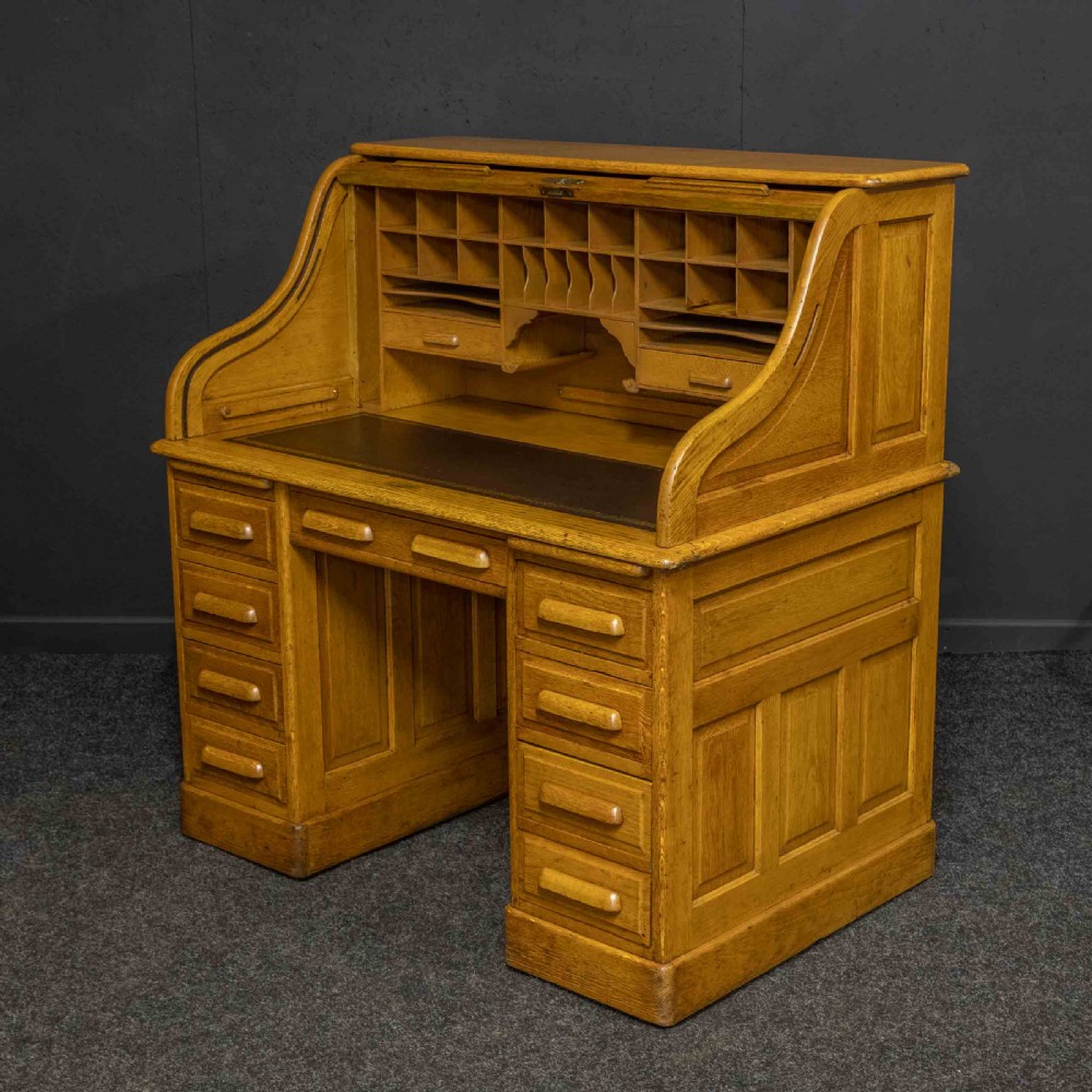 An early 20th-century golden oak roll top desk featuring open fitted pigeonhole compartments, a dark leather writing inset, and twin drawer pedestals. View IX