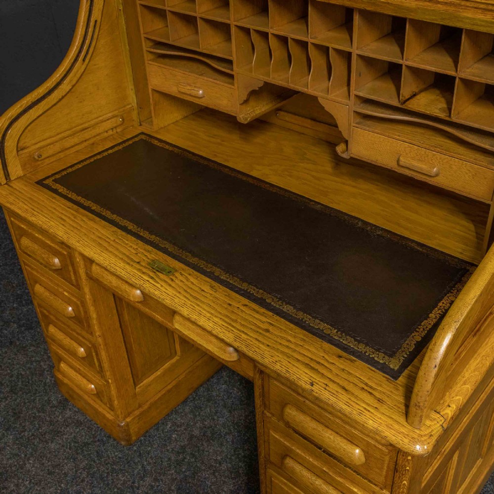 An early 20th-century golden oak roll top desk featuring open fitted pigeonhole compartments, a dark leather writing inset, and twin drawer pedestals. View X