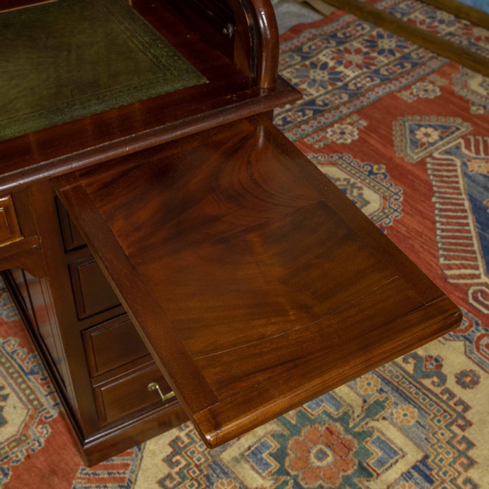 A mahogany roll top desk featuring a curved tambour shutter, internal pigeonhole compartments, a green leather inset writing surface, and twin pedestal drawers with brass drop handles. View V