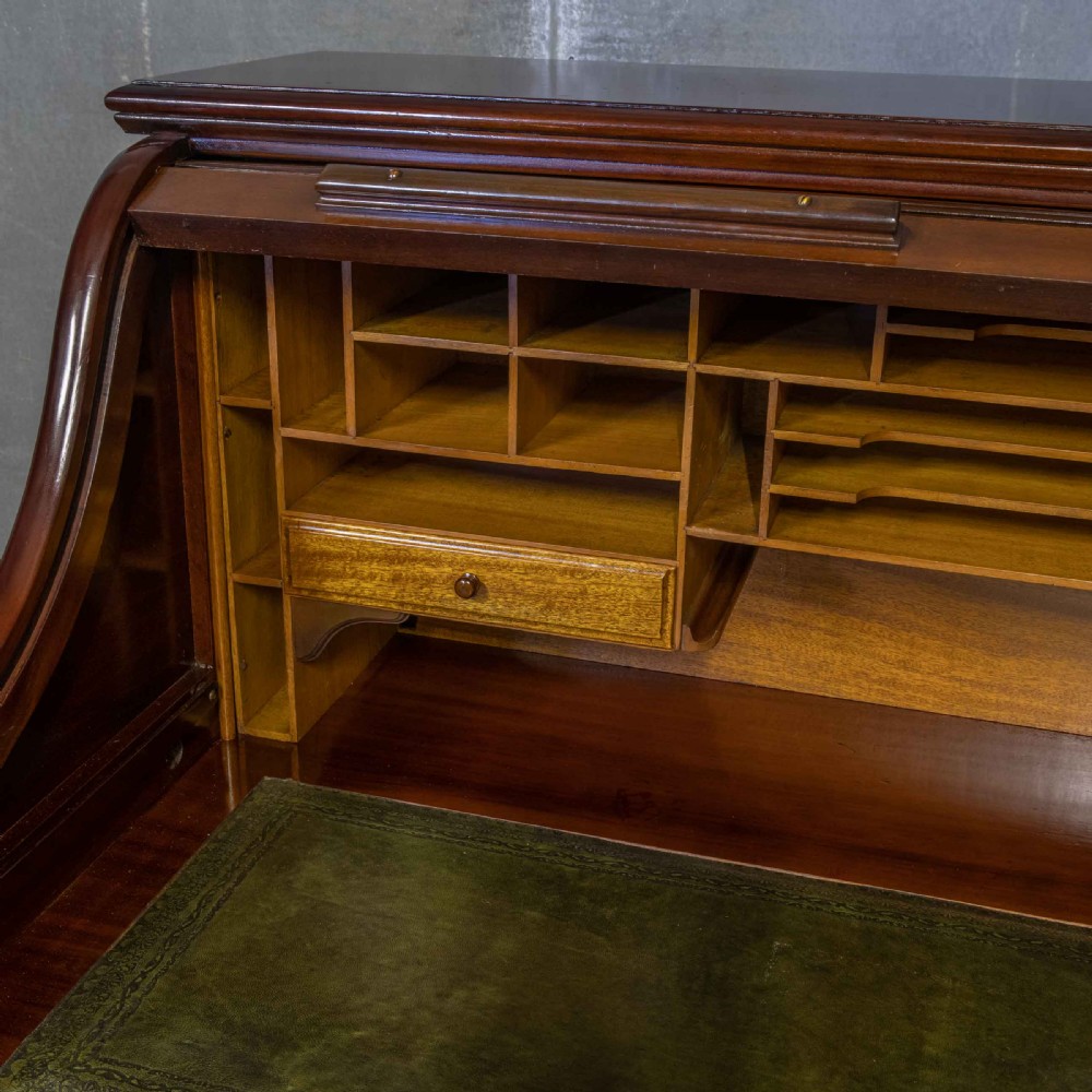 A mahogany roll top desk featuring a curved tambour shutter, internal pigeonhole compartments, a green leather inset writing surface, and twin pedestal drawers with brass drop handles. View VII