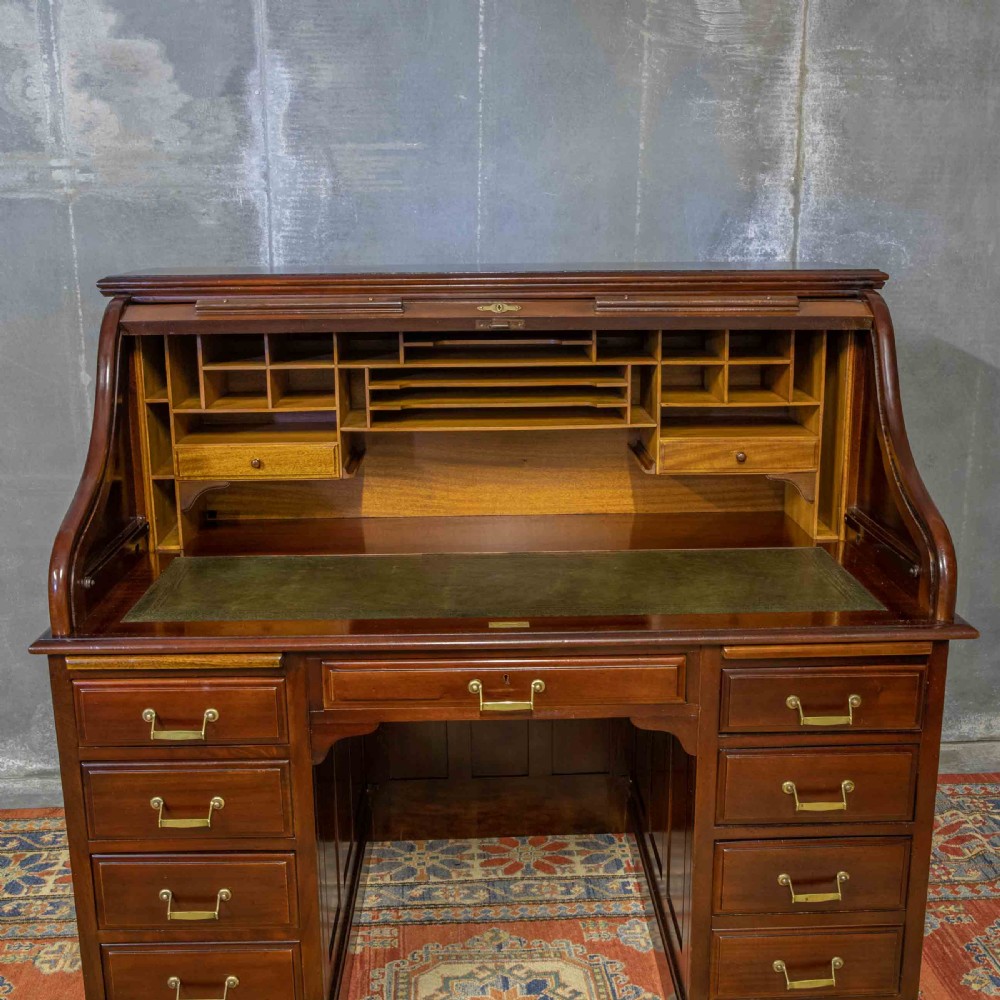 A mahogany roll top desk featuring a curved tambour shutter, internal pigeonhole compartments, a green leather inset writing surface, and twin pedestal drawers with brass drop handles. View IX