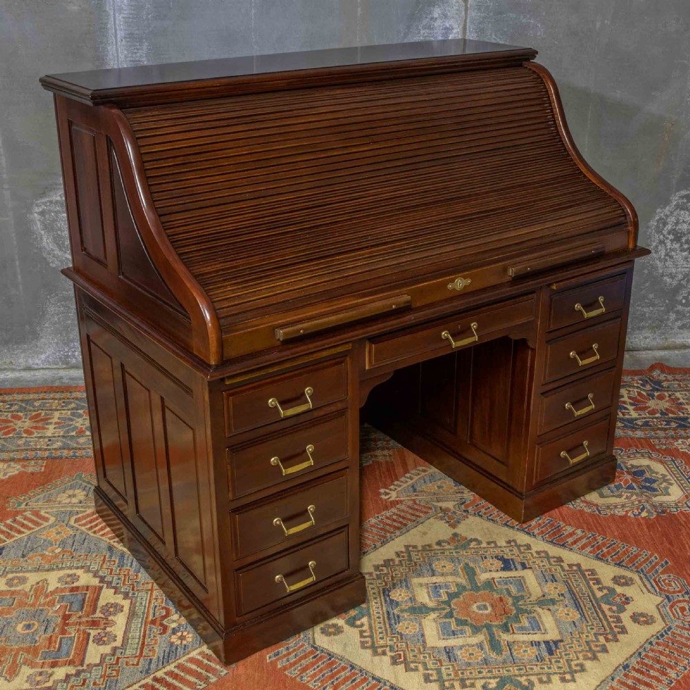 A mahogany roll top desk featuring a curved tambour shutter, internal pigeonhole compartments, a green leather inset writing surface, and twin pedestal drawers with brass drop handles. View XIV