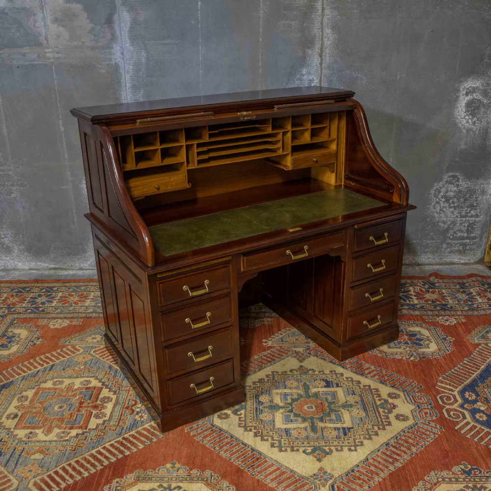 A mahogany roll top desk featuring a curved tambour shutter, internal pigeonhole compartments, a green leather inset writing surface, and twin pedestal drawers with brass drop handles. View XV