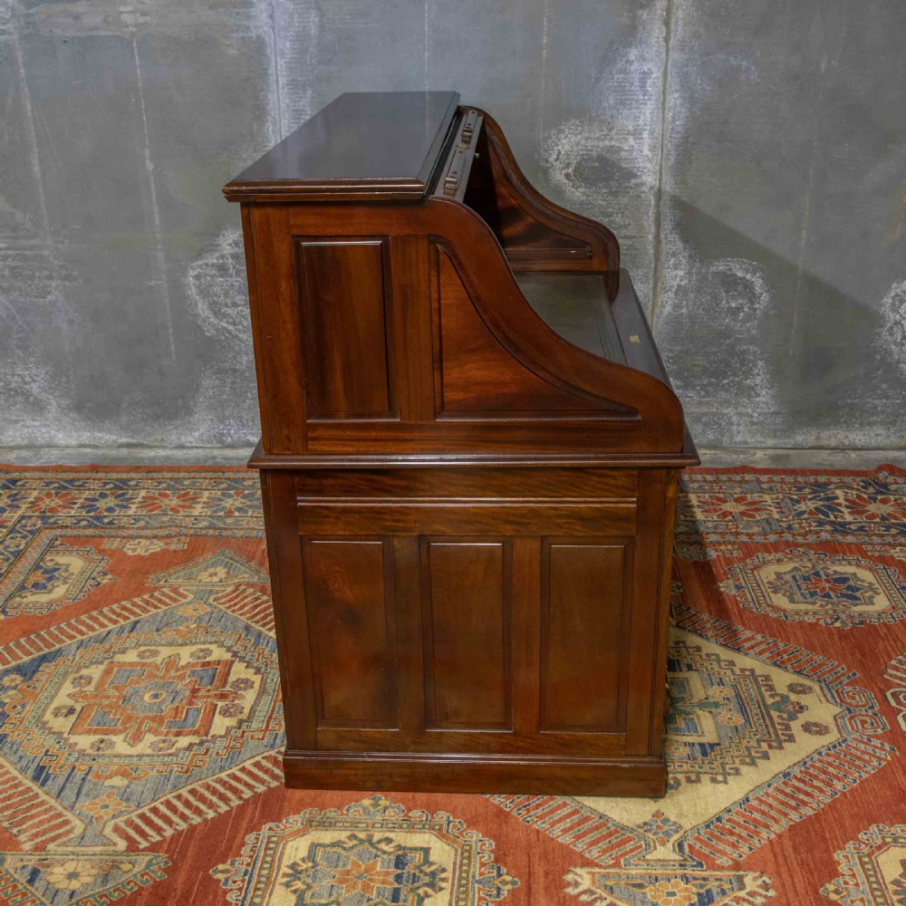 A mahogany roll top desk featuring a curved tambour shutter, internal pigeonhole compartments, a green leather inset writing surface, and twin pedestal drawers with brass drop handles. View XVI