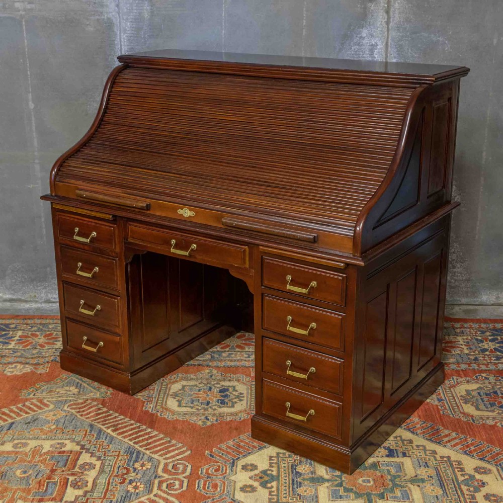 A mahogany roll top desk featuring a curved tambour shutter, internal pigeonhole compartments, a green leather inset writing surface, and twin pedestal drawers with brass drop handles. View XIX