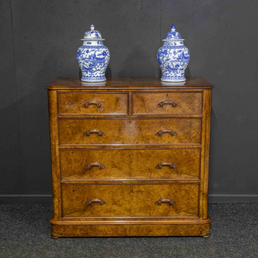 A Victorian burr walnut chest of drawers featuring a two-over-three drawer arrangement with carved handles and rounded corners on a plinth base. View V