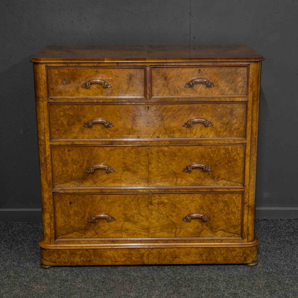 A Victorian burr walnut chest of drawers featuring a two-over-three drawer arrangement with carved handles and rounded corners on a plinth base. View VII