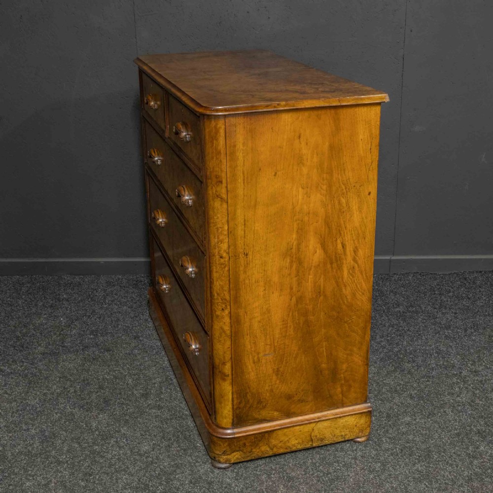 A Victorian burr walnut chest of drawers featuring a two-over-three drawer arrangement with carved handles and rounded corners on a plinth base. View IX