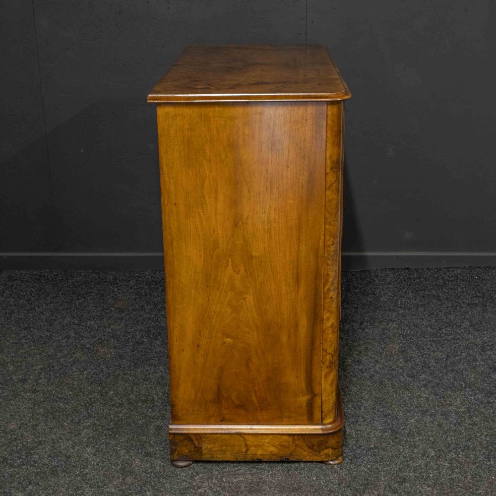 A Victorian burr walnut chest of drawers featuring a two-over-three drawer arrangement with carved handles and rounded corners on a plinth base. View XI
